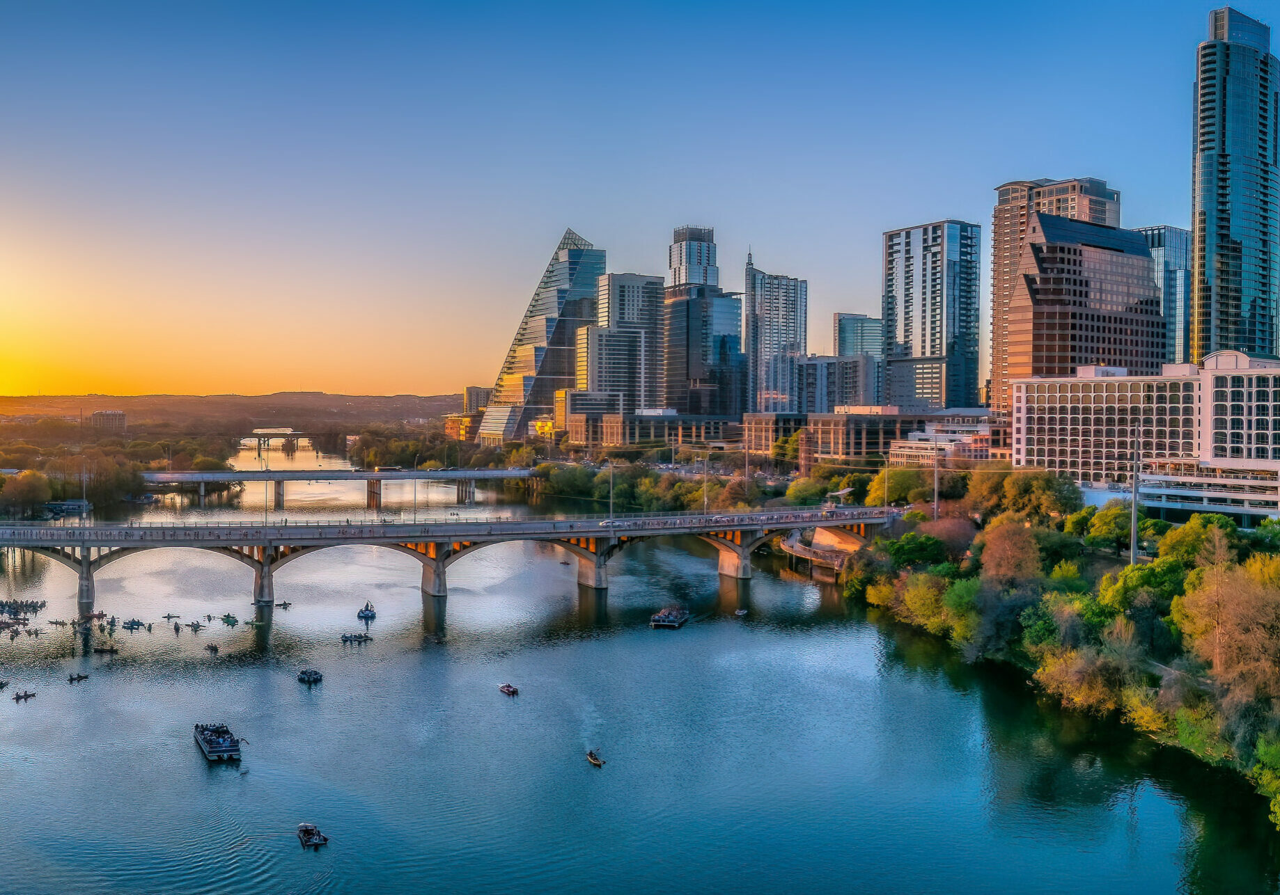 Austin, Texas- Cityscape against the sunset sky background. There are skycrapers on both sides with reflective glass walls and bridges over the Colorado river with boats.